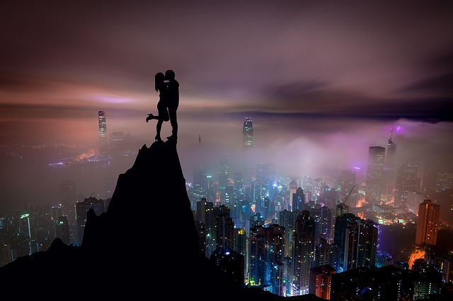 Muž a žena sa bozkávajú na vrchole kopca s panorámou rozsvieteného mesta za nimi.jpg